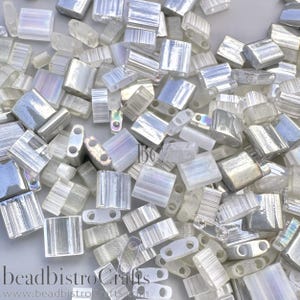 May include: A collection of rectangular, two-hole glass beads with an iridescent, semi-translucent white finish. The beads feature a ribbed texture and are scattered across the frame. The text beadbistroCrafts is visible at the bottom.