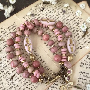 Peut inclure: Un bracelet multi-rangs compos&eacute; de perles roses et dor&eacute;es, avec un pendentif en forme de libellule et de croissant de lune. Le bracelet est pr&eacute;sent&eacute; sur un livre avec du texte sur les trag&eacute;dies de Leigh Hunt.