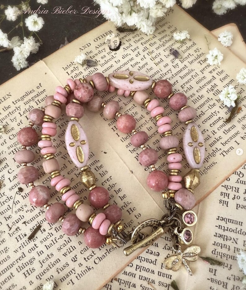 Peut inclure: Un bracelet multi-rangs compos&eacute; de perles roses et dor&eacute;es, avec un pendentif en forme de libellule et de croissant de lune. Le bracelet est pr&eacute;sent&eacute; sur un livre avec du texte sur les trag&eacute;dies de Leigh Hunt.
