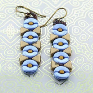 Könnte beinhalten: Ein Paar Hängeohrringe mit blauen und goldenen Perlen. Die Ohrringe bestehen aus kleinen, runden Perlen, die in einem sich wiederholenden Muster aufgereiht sind. Die Perlen sind hellblau mit einem metallischen Schimmer und werden mit goldenen Perlen in Form von kleinen Dreiecken durchsetzt.