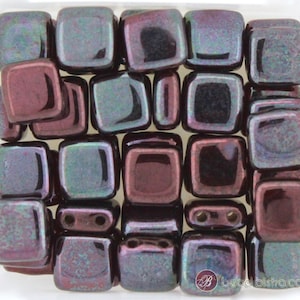May include: Square iridescent beads in shades of purple, teal, and black. The beads have a glossy finish and are arranged in a square container. Some beads have two holes for stringing. The beads are likely for jewelry making.