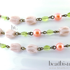 May include: Close-up of a delicate necklace featuring pale pink tulip-shaped beads, interspersed with green leaf-shaped beads and peach-colored pearl beads. The beads are strung on a bronze-colored chain. The necklace is displayed on a white surface.
