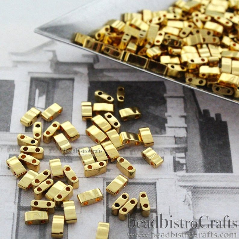 May include: Gold-colored metal beads with two holes each. The beads are arranged in a pile on a white and gray patterned surface. The beads are approximately 5mm in length.