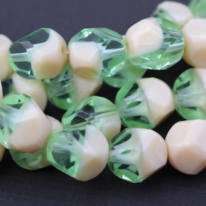May include: A collection of faceted beads featuring a dual-tone design with an opaque cream-coloured core encased in a translucent light green glass shell. The beads are arranged in a cluster, showcasing their geometric, multi-faceted surfaces.