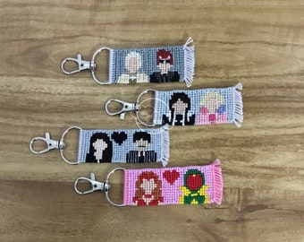May include: Handmade keychains with embroidered character designs. Each keychain features a rectangular design with a silver clasp. The characters are depicted in various colors, including gray, black, pink, red, and green. The keychains are displayed on a wooden surface.