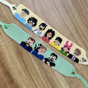 May include: Two friendship bracelets with colorful pixelated images of cartoon characters. The top bracelet is cream with a yellow, red, and blue design. The bottom bracelet is green with a blue, black, and white design.