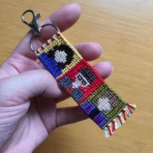 May include: A handmade, colorful keychain with a woven design. The keychain features a silver clasp and a rectangular woven section with a geometric pattern in red, blue, yellow, and brown. The edges have small fringe.