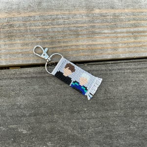 May include: A handmade keychain featuring a pixelated image of two figures. The keychain has a silver clasp and a gray, black, brown, blue, and white design. The keychain is made of woven material and has a fringed edge.