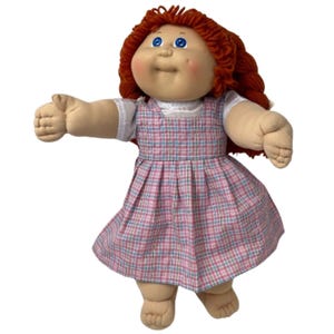 May include: A vintage Cabbage Patch Kid doll with red yarn hair, blue eyes, and rosy cheeks. The doll wears a white blouse and a pink and blue plaid dress. The doll's arms are outstretched, and it has a soft, plush body.