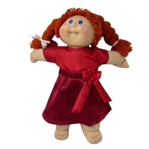 May include: A vintage doll with red yarn pigtails tied with white ribbons. The doll has blue eyes, rosy cheeks, and is wearing a red dress with a bow at the waist. The doll's arms are outstretched.