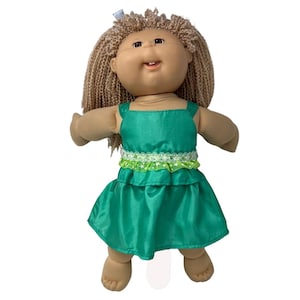 May include: A vintage doll with tan skin, brown eyes, and light brown, curly hair. The doll wears a green dress with a white and green trim around the waist. A small blue bow is in the doll's hair.
