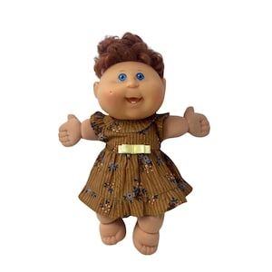 May include: A doll with brown yarn hair, blue eyes, and a dimpled face. The doll wears a brown dress featuring thin vertical stripes, a small floral pattern, a ruffled collar, and a pale yellow ribbon bow at the waist. The doll has outstretched arms.