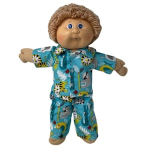 May include: A doll with a tan face, blue eyes, and light brown yarn hair. The doll is wearing a blue pajama set with a colorful jungle animal print, including tigers, sloths, and alligators. The pajamas have a button-down shirt and matching pants.