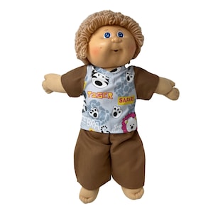 May include: A Cabbage Patch Kid doll wearing a safari-themed outfit. The doll has blonde yarn hair, blue eyes, and rosy cheeks. The outfit includes a brown shirt and pants, and a light blue vest with animal prints and the words "TIGER" and "SAFARI".