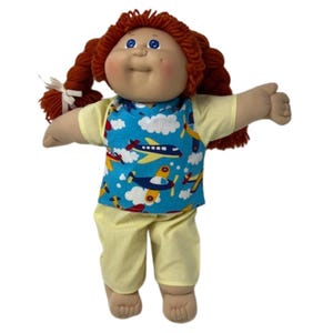 May include: A vintage doll with red yarn hair styled in pigtails, wearing a blue shirt with airplane graphics and yellow pants. The doll has a soft, fabric body and a smiling face with blue eyes.
