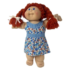 May include: A vintage doll with red yarn pigtails tied with white ribbons. The doll has blue eyes, freckles, and is wearing a blue and white floral print dress with a gingham bodice. The doll's arms are outstretched.