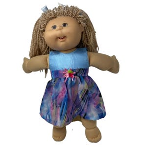 May include: A vintage doll with tan skin, blonde yarn hair, and a light blue dress top. The dress has a colorful, abstract pattern in shades of blue, purple, and pink. A pink flower with green leaves accents the waist. A blue ribbon is in the hair.