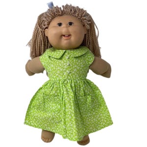 May include: A vintage doll with tan skin, brown eyes, and light brown yarn hair. The doll wears a green dress with a white floral pattern, a white collar, and two pockets. A small blue bow is in the doll's hair.