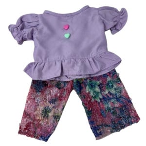 May include: A doll outfit featuring a lavender top with ruffled sleeves and hem, adorned with two heart-shaped buttons. The pants have a colorful floral pattern in shades of pink, blue, and green. This set is designed for dolls.
