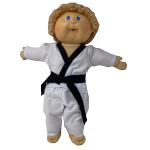May include: A Cabbage Patch Kid doll wearing a white karate uniform with a black belt. The doll has blonde, curly hair, blue eyes, and rosy cheeks. The doll's arms are outstretched.