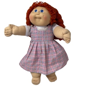 May include: A vintage Cabbage Patch Kid doll with red yarn hair and blue eyes. The doll is wearing a pink plaid dress with a white undershirt. The doll has a soft, plush body and is in a standing position with arms outstretched.