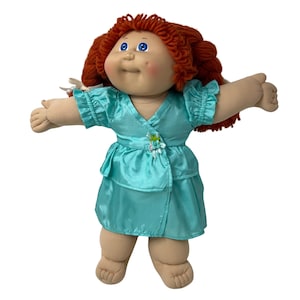 May include: A vintage doll with red yarn hair and blue eyes, wearing a turquoise satin dress. The doll's arms are outstretched, and it has a soft, plush body. The dress is adorned with a floral accent.