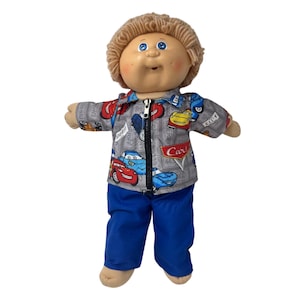 May include: A doll with light skin, rosy cheeks, and light brown yarn hair, wearing a gray zip-up jacket featuring a colorful Cars movie print with red, yellow, and blue vehicles, paired with solid blue pants. The doll has blue eyes and a soft body.