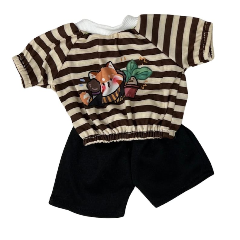 May include: A two-piece doll outfit featuring a brown and cream striped top with a cartoon fox graphic and black shorts. The top has short sleeves and a white neckline. The outfit is designed for dolls.