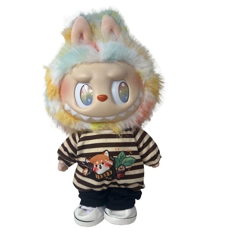 May include: A whimsical doll with a large, smiling face and colorful eyes. It wears a striped shirt with a fox graphic, black pants, and white sneakers. The doll's head is adorned with a fluffy, rainbow-colored hat with bunny ears.