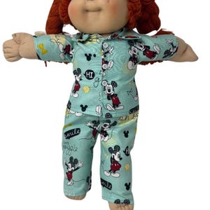 May include: A doll with yarn-like auburn hair and blue eyes wearing light blue pajamas featuring a repeating Mickey Mouse pattern, the text "HI", "smile", and "giggle", and small black mouse silhouettes. The pajamas have a button-down front.