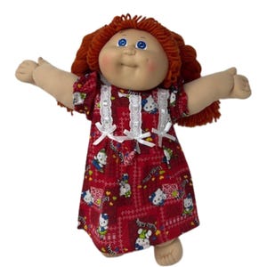 May include: A vintage Cabbage Patch Kid doll with red yarn hair and blue eyes. The doll is wearing a red dress with a white lace trim and white bows. The dress features a pattern of Hello Kitty characters.