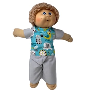 May include: A Cabbage Patch Kid doll wearing a light blue shirt with a jungle animal print and gray pants. The doll has brown curly hair, blue eyes, and rosy cheeks. The shirt features a pattern of elephants, birds, and other animals.
