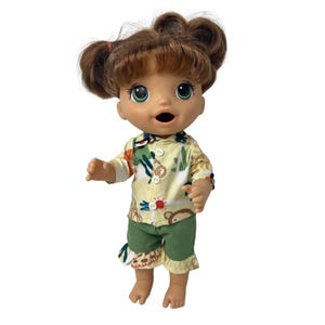 May include: A baby doll with brown hair styled in two buns and green eyes. The doll is wearing a yellow button-up shirt with a colorful print and green shorts with ruffled hems. The doll's mouth is open.