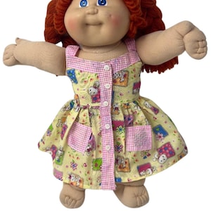 May include: A doll with yarn-like red hair, blue eyes, and a round face. It wears a yellow sleeveless dress featuring a pattern of small white rabbits and colorful flowers, accented with a pink and white checkered trim and four white buttons.