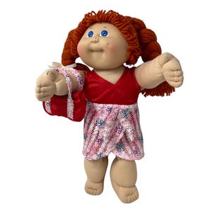 May include: A vintage doll with red yarn hair, blue eyes, and a rosy complexion. The doll wears a red satin top and a pink skirt with a floral pattern. It carries a red purse with white lace trim, a classic collectible.