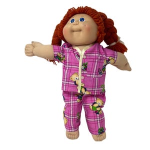 May include: A vintage doll with red yarn hair in pigtails, blue eyes, and rosy cheeks. The doll wears pink plaid pajamas with teddy bear and gift box patterns. The pajamas have a white trim and button-up shirt.