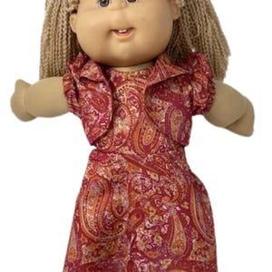 May include: A vintage Cabbage Patch Kid doll with blonde yarn hair and a light blue bow. The doll wears a red and orange paisley print dress with a matching jacket. The doll has a smiling expression and is in a standing position.