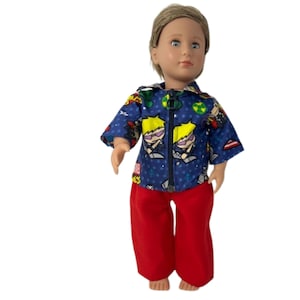 May include: A doll wearing a blue zip-up shirt with a superhero print and red pants. The shirt has a pattern of cartoon characters and stars. The doll has blonde hair and is standing against a white background.
