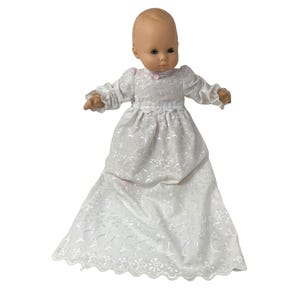 May include: A vintage baby doll with a bald head and brown eyes, dressed in a white eyelet gown. The gown features long sleeves, a small pink bow at the neckline, and a scalloped hem. The doll's arms are outstretched.