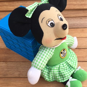 May include: A green and white gingham dress with a green bow and a Mickey Mouse logo patch. The plush toy has black ears and a brown face with black eyes and a red mouth.