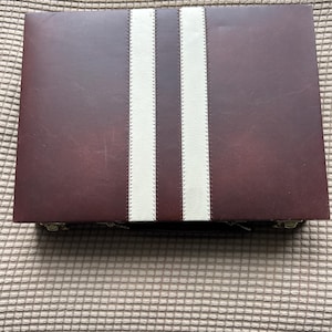 May include: A rectangular, burgundy-colored case with two vertical, cream-colored stripes down the center. The stripes are stitched onto the case. The case is closed and appears to be made of leather or a similar material.