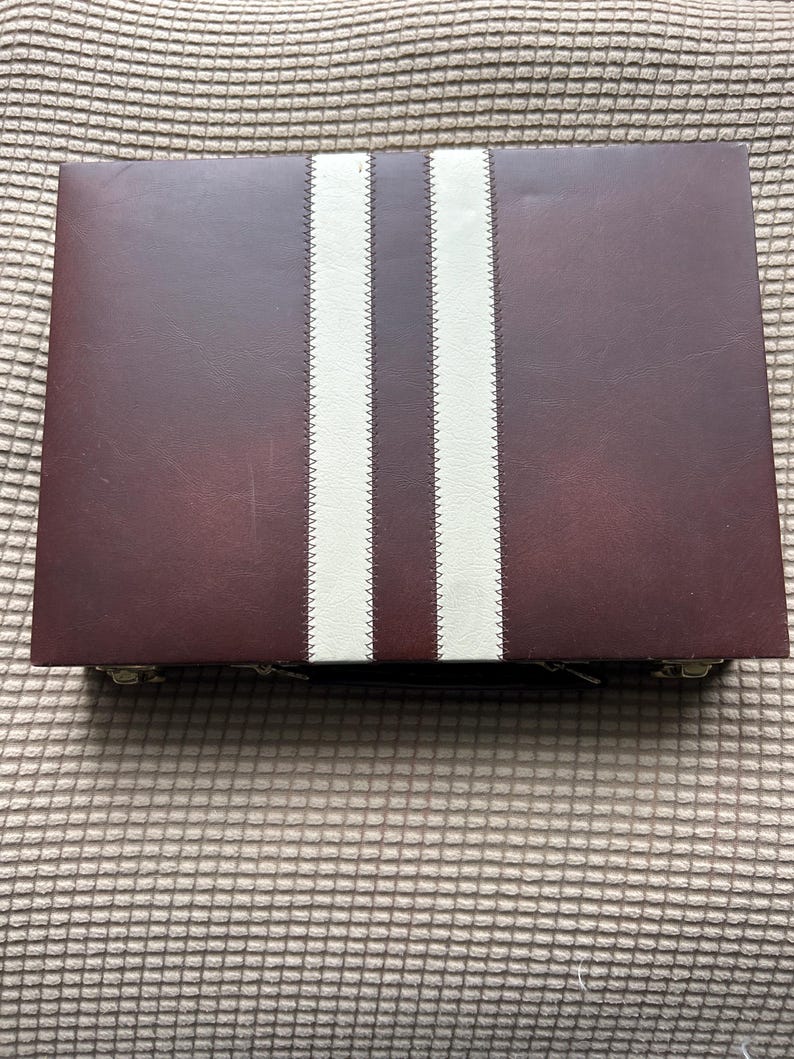 May include: A rectangular, burgundy-colored case with two vertical, cream-colored stripes down the center. The stripes are stitched onto the case. The case is closed and appears to be made of leather or a similar material.