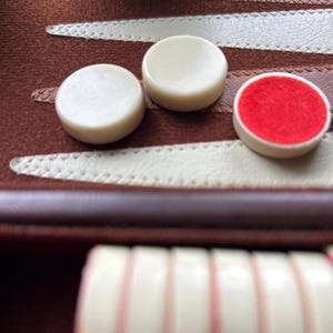 May include: Close-up of backgammon game pieces. The ivory-colored game pieces are round, with one piece featuring a red felt center. The game board is brown with white stitching.