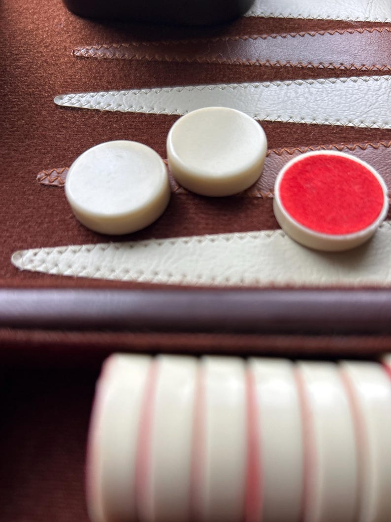 May include: Close-up of backgammon game pieces. The ivory-colored game pieces are round, with one piece featuring a red felt center. The game board is brown with white stitching.