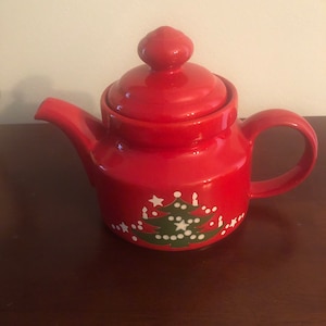 May include: Red ceramic teapot with a green Christmas tree design and white stars and dots. The teapot has a lid with a knob on top.