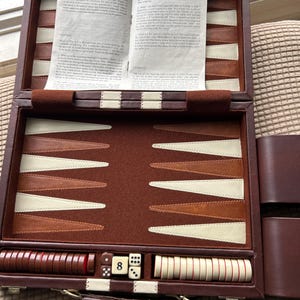 May include: Open brown leather backgammon set with ivory and brown playing pieces, dice, and a doubling cube showing the number 8. The game board is open, revealing the playing surface and instructions.