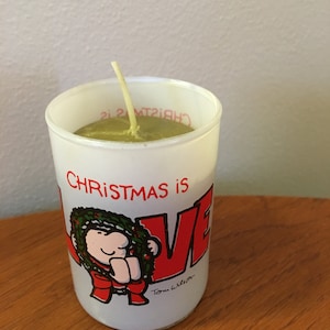May include: A white votive candle holder with a green and red illustration of a cartoon character holding a wreath. The text "Christmas is LOVE" is printed on the holder. The candle is green and has a white wick.