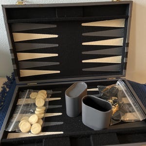 May include: A gray backgammon set with a black interior, cream and gray triangles, and a black felt playing surface. Includes cream-colored game pieces in a plastic bag, dice, and two gray dice cups.
