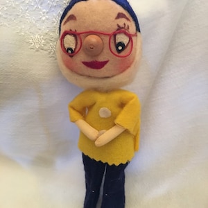 May include: A vintage felt doll with a blue hat, red glasses, and a yellow dress. The doll has a round face with a button nose and painted features.