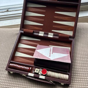 May include: Open backgammon set with a brown leather case, featuring a playing surface with alternating brown and cream-colored points. Includes dice, checkers, and a Backgammon instruction card.